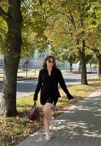 Μαύρο κομμένο σακάκι συνδυασμένο με μαύρο μίνι φόρεμα με πλάγιο άνοιγμα. Ασημί ψηλοτάκουνα και τσάντα σε χρώμα μπορντό. Φωτεινός ηλιόλουστος χώρος πάρκου.