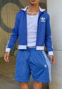 Giacca da tuta blu Adidas con accenti e logo bianchi, abbinata a pantaloni corti blu coordinati con tre strisce bianche ai lati.
