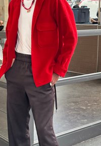 Personne portant une veste rouge sur une chemise blanche avec un collier de perles rouges et un pantalon noir, debout avec une main dans la poche.