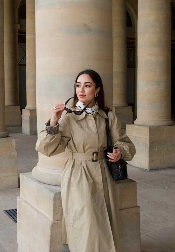 Mujer con gabardina beige que sostiene gafas de sol y bolso negro, de pie junto a grandes columnas de piedra en un entorno arquitectónico clásico.