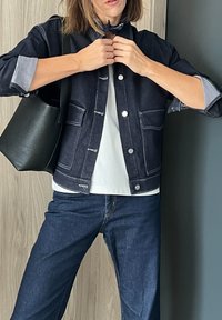 Τζάκετ από τζην σε σκούρο μπλε με ασημί κουμπιά και άσπρες ραφές, φορεμένο με λευκή μπλούζα και σκούρα μπλε τζιν. Μαύρη τσάντα ώμου.