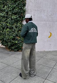 Green sweatshirt featuring a "QUOTELL HEIGHTS" graphic on the back, matched with loose, faded grey trousers and black shoes. The wearer is standing against an ivy-covered wall.