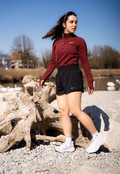 Camiseta deportiva de manga larga color burdeos con cuello con cremallera, combinada con pantalones cortos negros. Zapatillas blancas con acentos azules. Entorno natural con madera flotante.