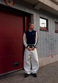 Homme aux cheveux coiffés en arrière portant une veste bleu marine, blanche et rouge, un pantalon large blanc et des chaussures noires, se tenant contre un mur carrelé et une porte rouge.