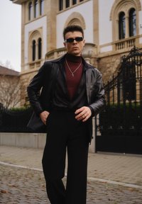 Zwarte leren jas over een bordeauxrode coltrui, gecombineerd met zwarte wijde broek. Accessoires zijn onder andere een zonnebril en een kruis-ketting.