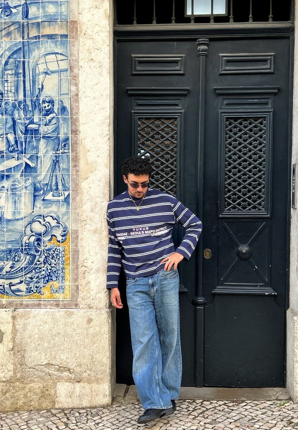 Moški z sončnimi očali in črtasto srajco se naslanja na steno ob okrašena črna vrata in modro keramično fresko, ki prikazuje ljudi in stroje.