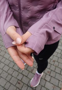Chaqueta acolchada lilac con un patrón texturizado y bumpy; mangas ajustadas; manos entrelazadas; persona vistiendo leggings negros y zapatillas en un tono púrpura claro.