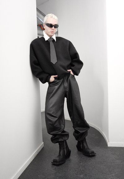 Persona con cabello corto y rubio y gafas de sol apoyada contra una pared blanca, vistiendo un suéter negro de gran tamaño, pantalones anchos de cuero, botas negras y una corbata negra.