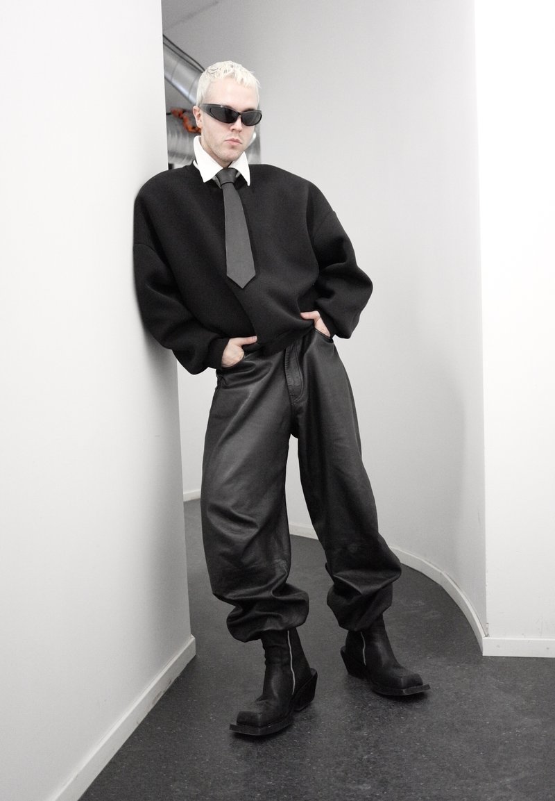 Personne aux cheveux blonds courts et lunettes de soleil, appuyée contre un mur blanc, portant un pull noir surdimensionné, un pantalon large en cuir, des bottes noires et une cravate noire.