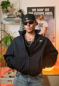 Veste noire zippée avec un logo Nike, portée sur un t-shirt noir, assortie à un jean bleu. Accessoires incluent des chaînes et des lunettes de soleil.