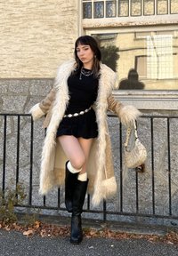 Een tan jas met faux fur-kraag over een zwarte jurk, gecombineerd met kniehoge zwarte laarzen en witte kniekousen, terwijl ze een kleine geweven handtas vasthoudt.