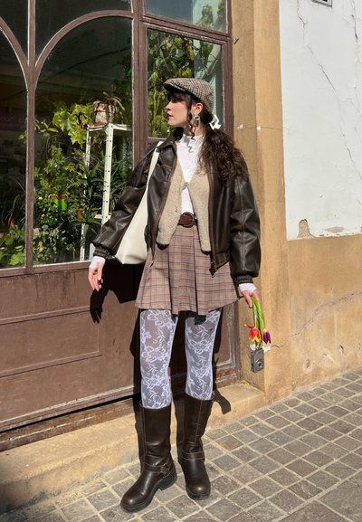 Mujer de pie fuera de un edificio con una chaqueta de cuero marrón, falda de cuadros, medias blancas de encaje y botas, sosteniendo tulipanes cerca de una puerta de cristal.