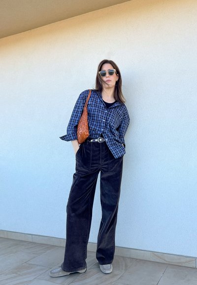 Camisa de cuadros en negro y azul, pantalones de pana anchos en negro, zapatos grises y un bolso tejido marrón. Las gafas de sol redondas completan el look.