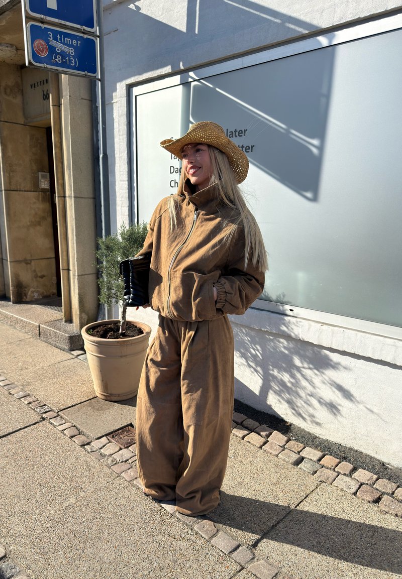 Jeune femme portant une tenue marron et un chapeau de paille, debout sur un trottoir en ville près d'une plante en pot et d'un panneau de stationnement.