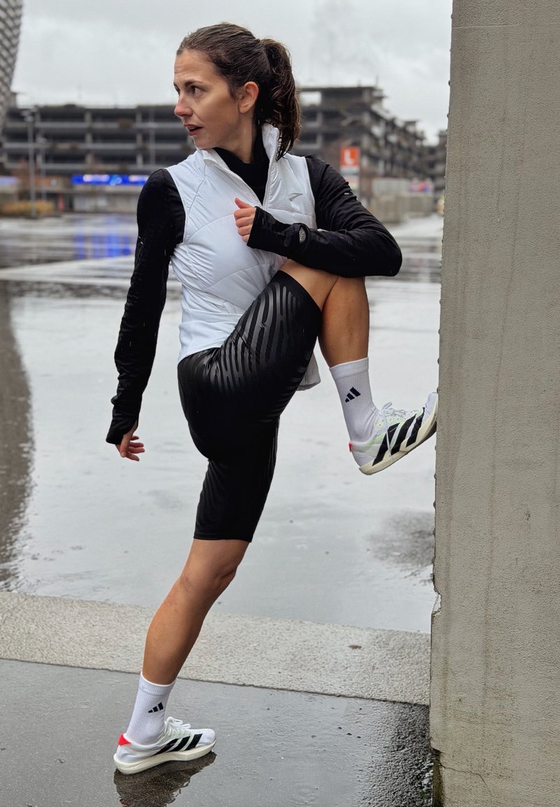 Woman in sportswear stretching one leg against a wall on a wet urban street under overcast sky.