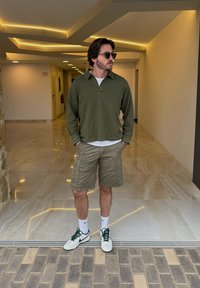 Maglione a maniche lunghe verde oliva, pantaloni cargo beige, calzini bianchi e sneakers con accenti verdi, in piedi in uno spazio interno moderno con pavimenti in marmo.