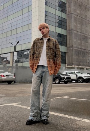 Joven con cabello teñido claro que lleva una camisa de cuadros marrón y naranja sobre una camiseta blanca, jeans holgados y zapatos negros, de pie en un estacionamiento urbano.