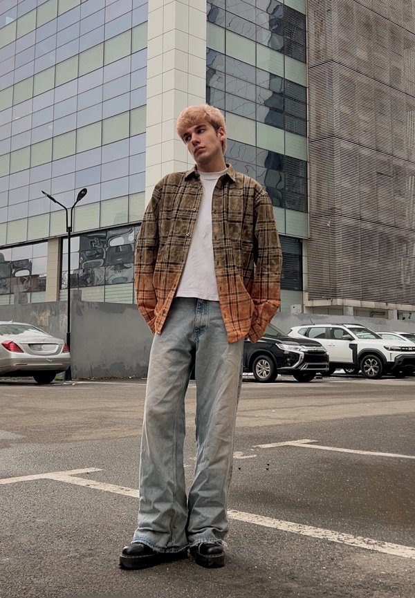 Ung mann med blondt, farget hår iført brun og oransje rutete skjorte over hvit t-skjorte, løse jeans og svarte sko, stående på en urban parkeringsplass.