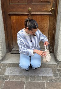 Ένα άτομο με ριγέ μακρυμάνικη μπλούζα γονατίζει σε λιθόστρωτο δρόμο, κρατώντας μια ανοιχτόχρωμη βαλίτσα με κορδόνι.