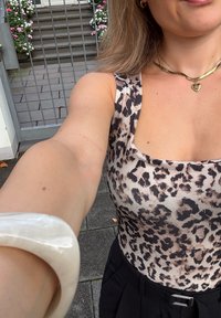 Beige und schwarze, enganliegende Leopardenmuster-Bodysuit mit breiten Trägern. Weißes, klobiges Armband am linken Arm. Schwarze, hoch geschnittene Hosen sichtbar.