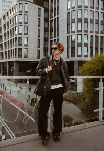 Chaqueta de cuero negra sobre un suéter de punto oscuro, pantalones negros sueltos y gafas de sol. Sosteniendo una bolsa de tela al hombro, de pie sobre un puente.