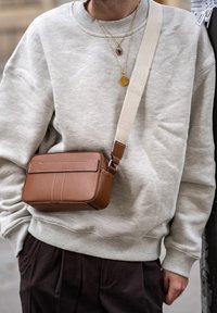 Brun lærcrossbody-veske med hvite sømmer, rektangulær form og justerbar kanvasstropp, båret over en grå hettegenser.