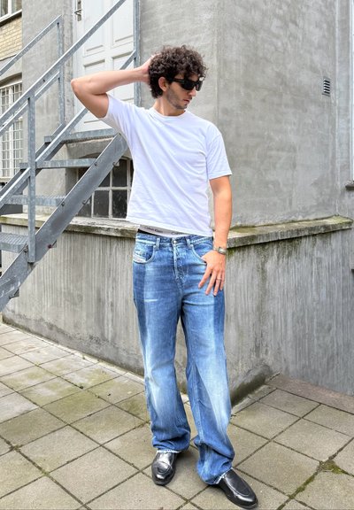 Camiseta blanca, jeans azul claro de corte holgado con un aspecto desgastado, gafas de sol negras y zapatos negros. El modelo está apoyado contra una pared de concreto.