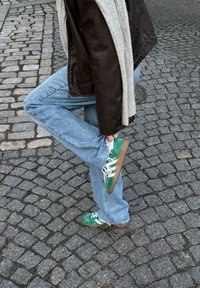 Groene en witte sneakers met een suède textuur, bruine zolen, gestreepte accenten, gedragen met lichtblauwe jeans en een bruine jas. Achtergrond van kasseien.