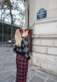 Black and grey argyle sweater paired with a red and black tartan maxi skirt, complemented by a flowing blonde ponytail and a red hair ribbon.