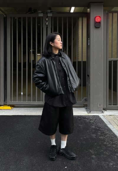 Mujer con chaqueta de cuero negro y pantalones cortos se encuentra con las manos en los bolsillos cerca de una puerta cerrada y un semáforo rojo.