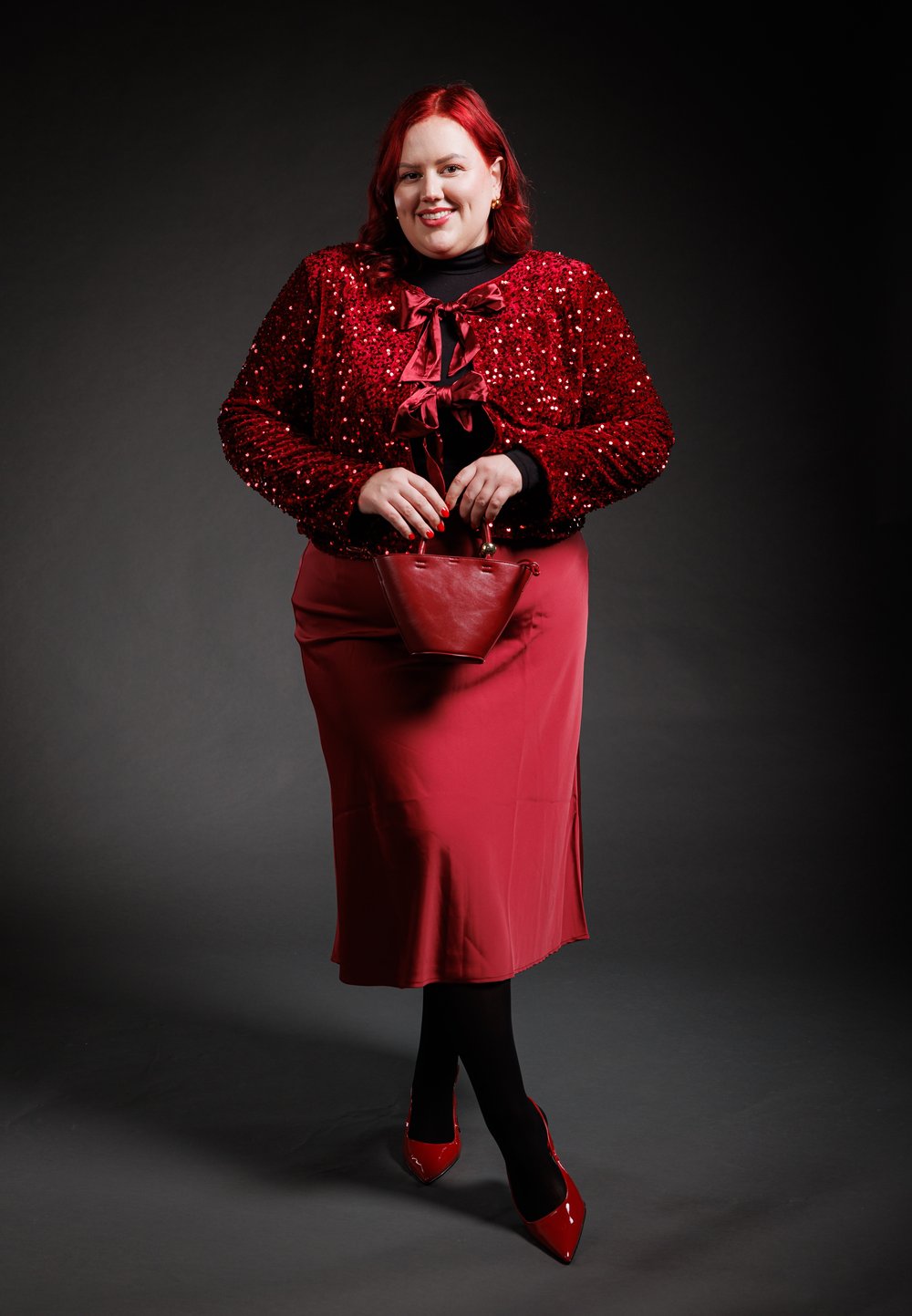 Veste rouge à sequins avec détail nœud, jupe en satin rouge, collants noirs et escarpins rouges vernis. Petit sac à main rouge complète la tenue.