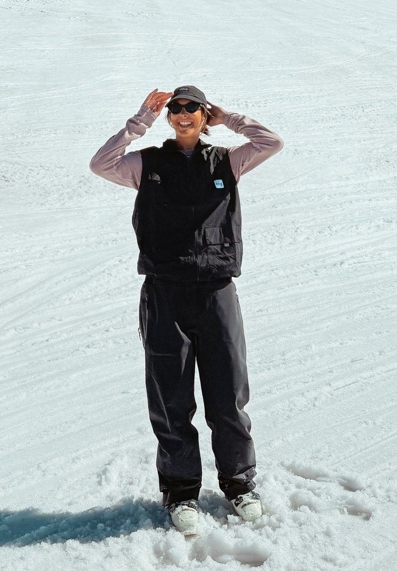 Gilet nero sopra una camicia a maniche lunghe color lavanda, pantaloni neri e stivali da sci bianchi. Gli accessori includono occhiali da sole e un cappellino scuro. Sfondo innevato.