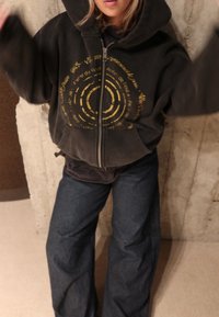 Felpa nera con zip e design circolare giallo, abbinata a jeans di denim scuro a gamba larga. Il tessuto sembra morbido con una vestibilità comoda.