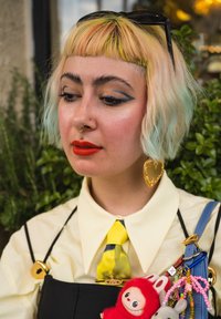 Joven con cabello corto y multicolor, delineador de ojos azul intenso, pintalabios rojo, perforaciones en el septum y el labio de oro, grandes pendientes en forma de corazón dorado y accesorios coloridos.