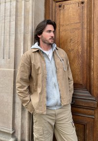 Καφέ βαμβακερό μπουφάν πάνω από γκρι φούτερ, συνδυασμένο με χακί cargo παντελόνι. Λείες υφές, άνετος σχεδιασμός και λεπτομέρεια με μπροστινό φερμουάρ.