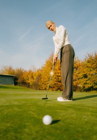 Golf-Putter mit schwarzem Kopf, silbernem Schaft; trägt weißen Langarmpullover, graue gestreifte Hose und weiße Turnschuhe; puttet auf grünem Gras.