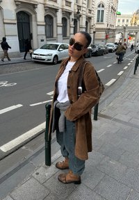 Femme portant un manteau en daim marron, une chemise blanche et un jean, debout sur un trottoir en ville près de voitures garées et de cyclistes dans une rue bordée de bâtiments historiques.