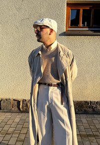 Trench coat bege claro sobre um suéter bege, calças creme e um cinto de couro castanho. Um boné cinza e óculos completam o visual.