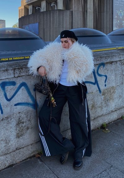Chaqueta de pelo sintético blanca y esponjosa, gorro negro con gráfico, camiseta blanca, pantalones de chándal negros con rayas blancas y zapatos negros con cordones.