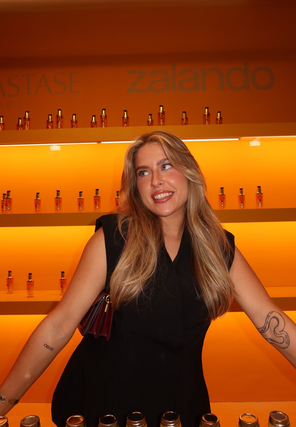 Hyllor som visar amberflaskor med hårprodukter mot en orange bakgrund. En person i en svart ärmlös topp poserar med en mörk handväska.