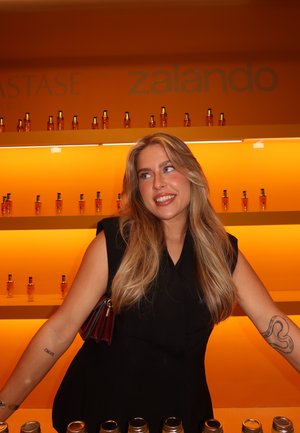 Estantes que exhiben botellas ámbar de productos para el cabello sobre un fondo naranja. Una persona con una camiseta sin mangas negra posa con un bolso oscuro.