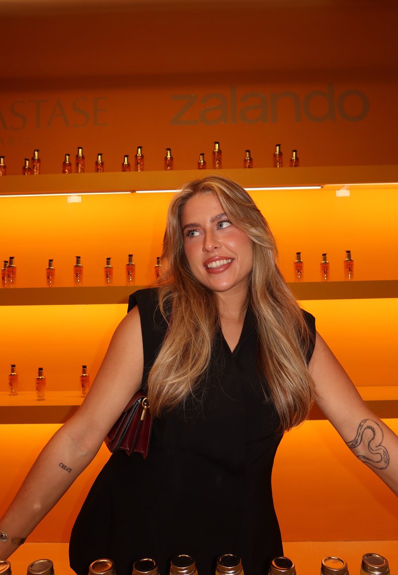 Planken met amberkleurige flessen haarproducten tegen een oranje achtergrond. Een persoon in een zwart mouwloos topje poseert met een donkere handtas.