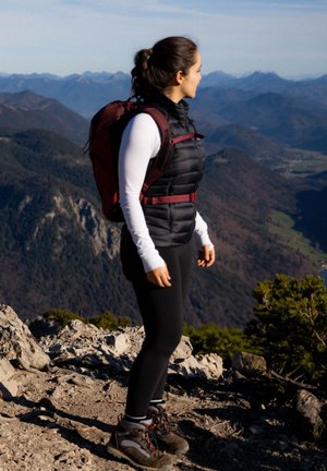 Gilet imbottito nero sopra una maglietta a maniche lunghe bianca, leggings neri e scarponi da escursionismo. Uno zaino bordeaux poggia sulla sua schiena tra un terreno roccioso.