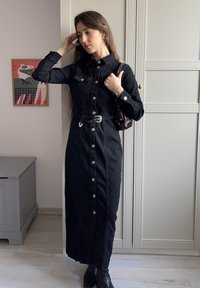 Juoda džinsinė suknelė marškinėliai su ilgomis rankovėmis, priekiniais sagais ir diržu. Nešiojama su tamsia rankine ir juodais aulinukais.