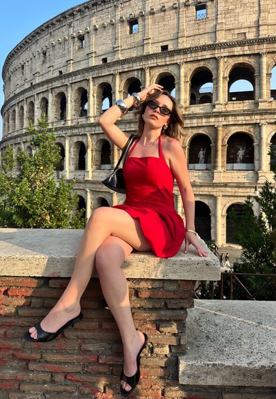 Vestido rojo sin mangas con un corsé ajustado, mules negras de punta abierta y gafas de sol. El Coliseo Romano es visible en el fondo.