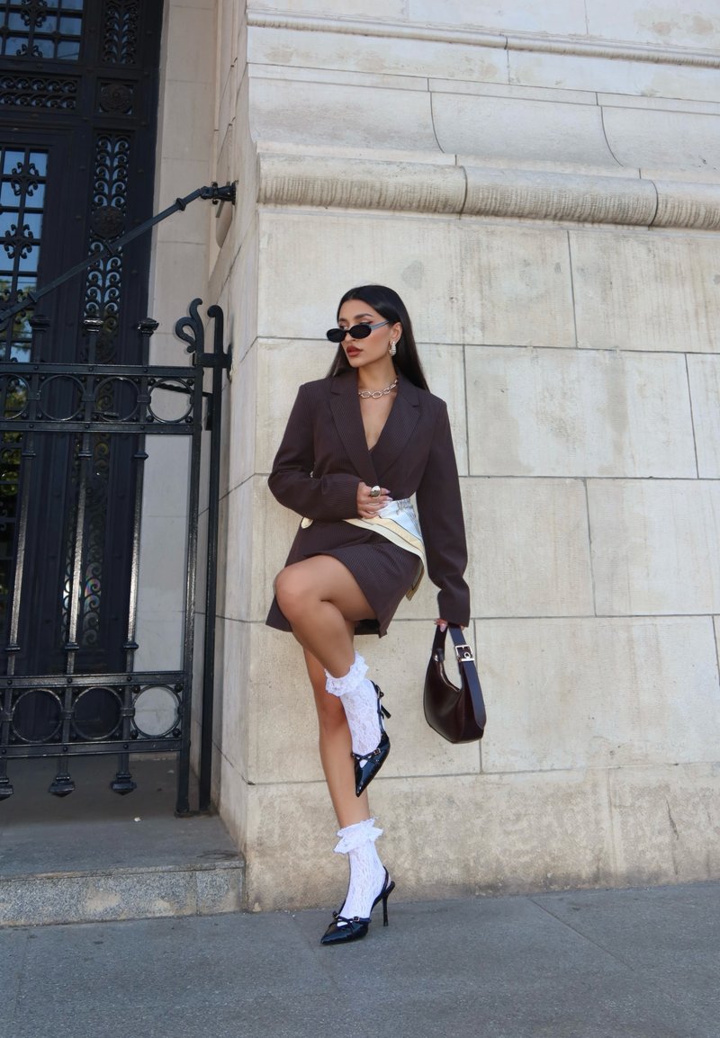 Brown pinstripe blazer over a white top, paired with a short skirt, lace white socks, and black heels, complemented by a dark handbag.