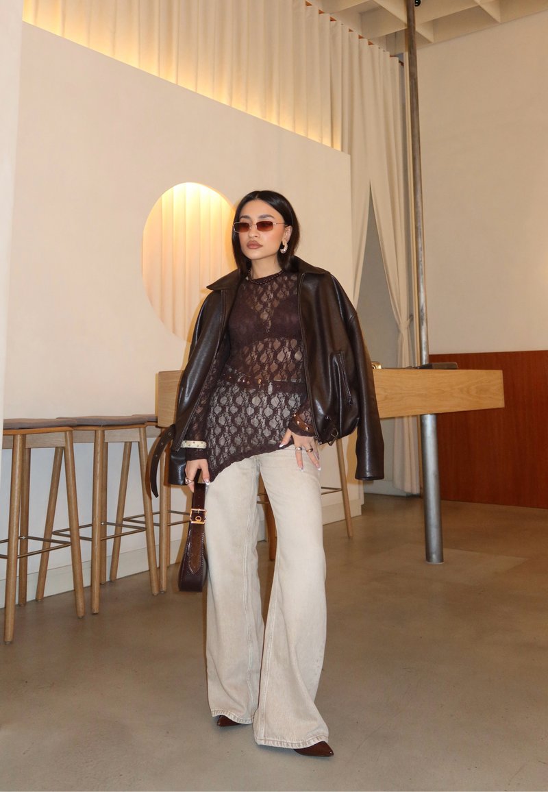 Naine, kes kannab päikeseprille, musta pitsist topi, tumedat nahktagi õlgadel, beeži laia lõikega pükse ja hoiab käes tumedat käekotti moodsas kohvikus.