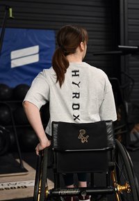 Grijs shirt met korte mouwen met 'HYROX' verticaal gedrukt op de achterkant, gecombineerd met een zwarte rolstoel met gouden accenten en meshdetails.