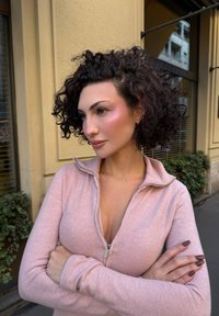 Jasnoróżowy sweter zapinany na zamek z miękką fakturą, z wysokim kołnierzem i długimi rękawami. Modelka ma kręcone włosy i skrzyżowane ramiona.