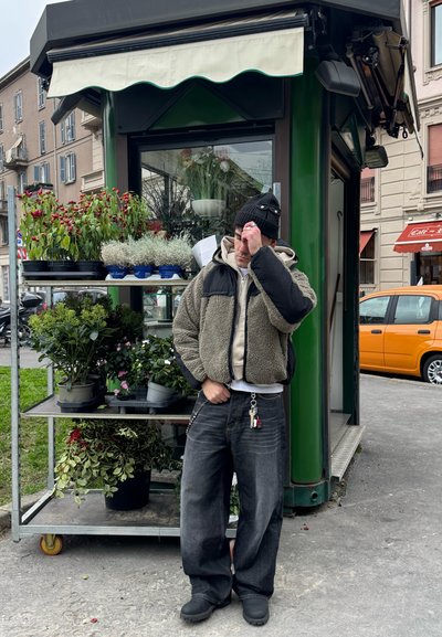 Chaqueta verde y negra difusa, sudadera beige, jeans oscuros. Puesto de flores con varias plantas en macetas y flores rojas en macetas brillantes.